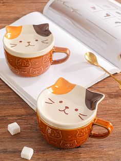 two coffee mugs with cats on them sitting on a table next to an open book