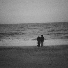 two people are standing on the beach looking out at the ocean in black and white