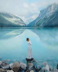 a woman standing on rocks in front of a body of water with mountains in the background