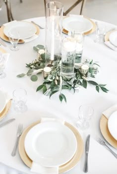 the table is set with white and gold plates, silverware, candles and greenery