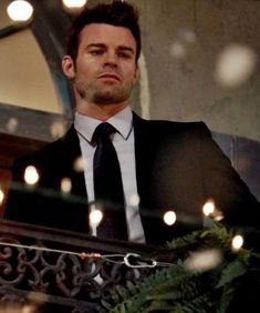 a man wearing a suit and tie standing in front of a balcony with christmas lights