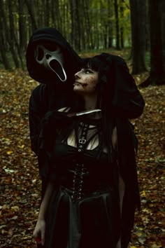 a woman in a black dress with a mask on her head is standing in the woods