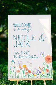 a welcome sign with flowers painted on it in front of some bushes and trees,