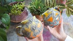 two people holding up vases with plants in them on a table next to potted plants