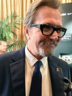 an older man wearing glasses and a suit smiling at the camera while standing next to a plant