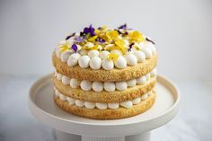 a three layer cake with white frosting and purple flowers on top is sitting on a plate