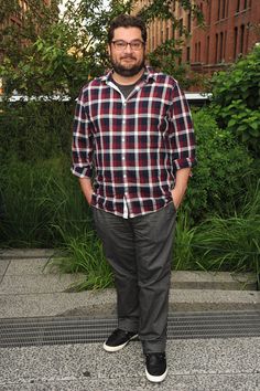 a man standing in front of a building with his hands in his pockets and wearing glasses