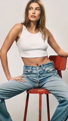 a woman sitting on top of a wooden chair next to a white wall and wearing jeans