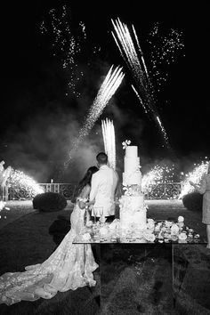 a newly married couple standing in front of their wedding cake at night with fireworks behind them