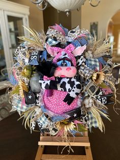 a cow wreath made out of fabric and other items on a wooden stand with a chandelier in the background