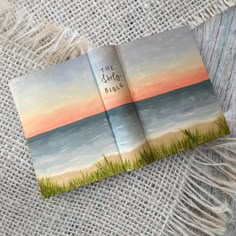 an open book sitting on top of a table next to some grass and the ocean