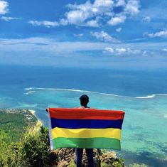 a man standing on top of a mountain holding a rainbow colored flag in front of the ocean