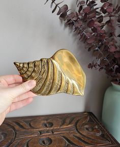 a person is holding a gold object in front of a plant and vase on a table