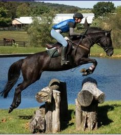 a man riding on the back of a black horse jumping over a wooden fence next to a lake