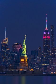 the statue of liberty is lit up at night in front of the city skyline and skyscrapers