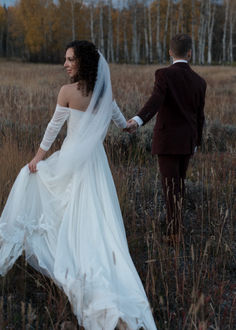 a bride and groom are walking through tall grass in their wedding attire, holding hands