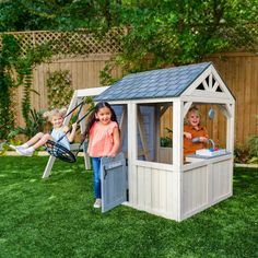 three children playing in a small wooden play house on green grass with trees and fence behind them