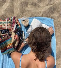 a woman laying on top of a blue towel next to a bag and book in the sand