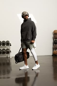 a man in grey hoodie and white shorts holding a black bag while standing next to a gym equipment rack