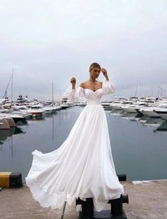 a woman in a long white dress standing on a dock next to some water and boats