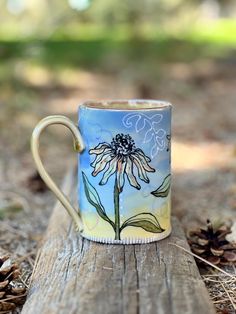 a painted coffee cup sitting on top of a wooden table next to pine cones and leaves