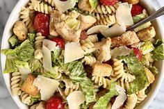 a white bowl filled with pasta salad on top of a marble counter topped with chicken, lettuce and cherry tomatoes