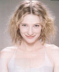 a woman with blonde hair wearing a white tank top and smiling at the camera while standing in front of a gray background