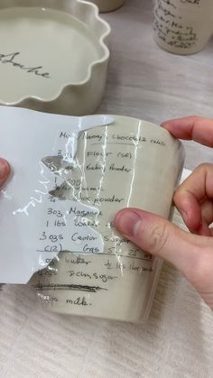 a person holding a piece of paper with writing on it next to cups and saucers