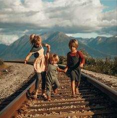 three children walking on train tracks in the mountains, one holding hands with the other