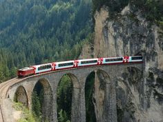 a red and white train traveling over a bridge next to a lush green forest covered hillside