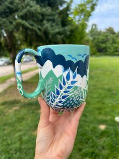 a hand holding up a painted coffee cup in the grass with trees and clouds on it