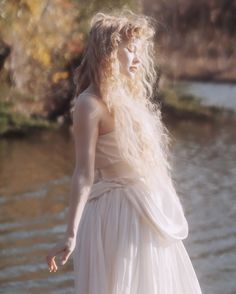 a woman with long hair standing in front of water wearing a white dress and holding her hand out to the side