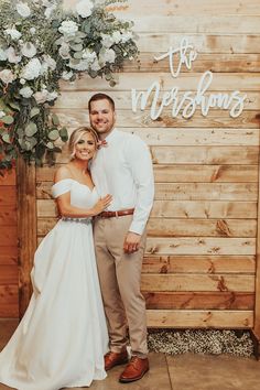 a bride and groom pose for a photo in front of a wooden wall decorated with flowers