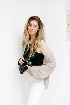 a woman is holding a camera and posing for a photo in front of a white wall