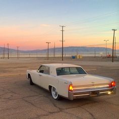 an old white car parked in a parking lot at sunset with power lines and mountains in the background