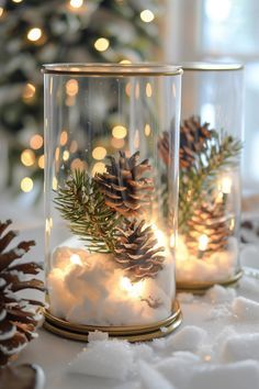 two glass jars filled with snow and pine cones are sitting on a table next to a christmas tree