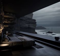 a bed sitting next to a large window near the ocean in front of a cliff