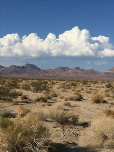 a dirt field with mountains and clouds in the sky behind it on a sunny day