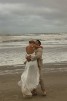 a man and woman hug on the beach as they walk towards the ocean with waves in the background