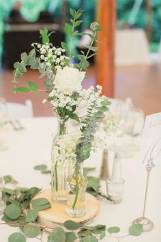 a vase filled with white flowers and greenery on top of a wooden tablecloth