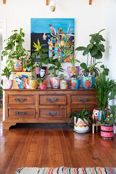 a wooden dresser topped with lots of potted plants next to a painting on the wall