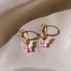 a pair of pink butterfly shaped earrings on a white cloth with gold hoops and an earring