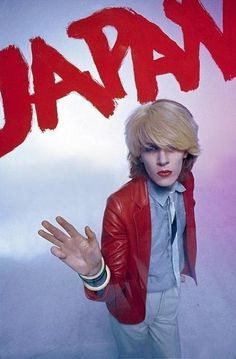 a man with blonde hair wearing a red jacket and tie standing in front of a poster