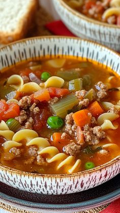 a bowl of soup with noodles, meat and vegetables on a red place mat next to bread