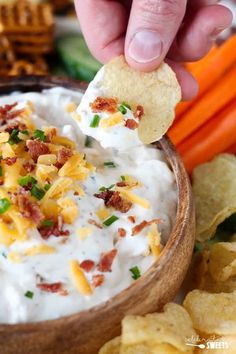 a hand holding a cracker over a bowl of dip with bacon, cheese and chives