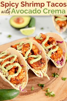 three shrimp tacos with avocado sauce on a wooden cutting board next to an avocado