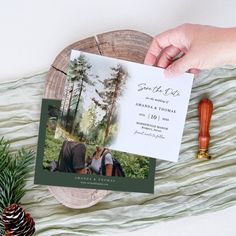 a person holding up a wedding card next to a pine cone and fir tree branch