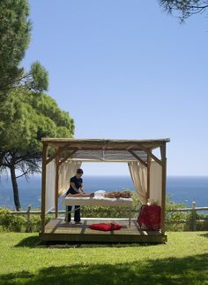 a man sitting on top of a bed under a canopy next to the ocean and trees