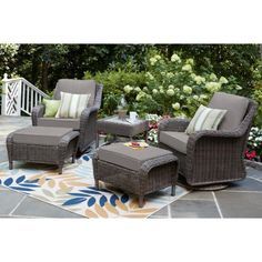 an outdoor patio with wicker furniture and flowers in the background, including two chairs and a footstool