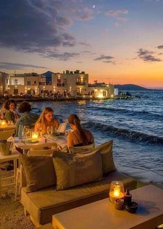 people are sitting at tables on the beach by the water with candles in their hands